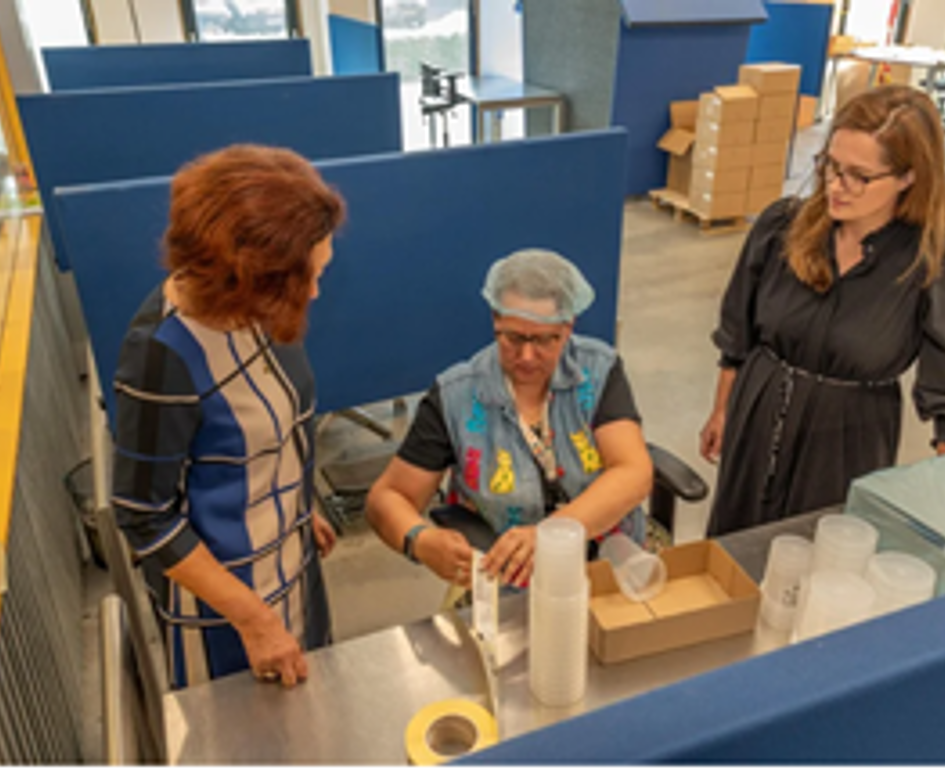 Algemeen directeur Marjolijn Dölle (links) en Daphne Siebenlist (teamleider beleid en communicatie) kijken op de werkvloer van WerkSaam Westfriesland mee over de schouder van medewerksters Olga Celighen   