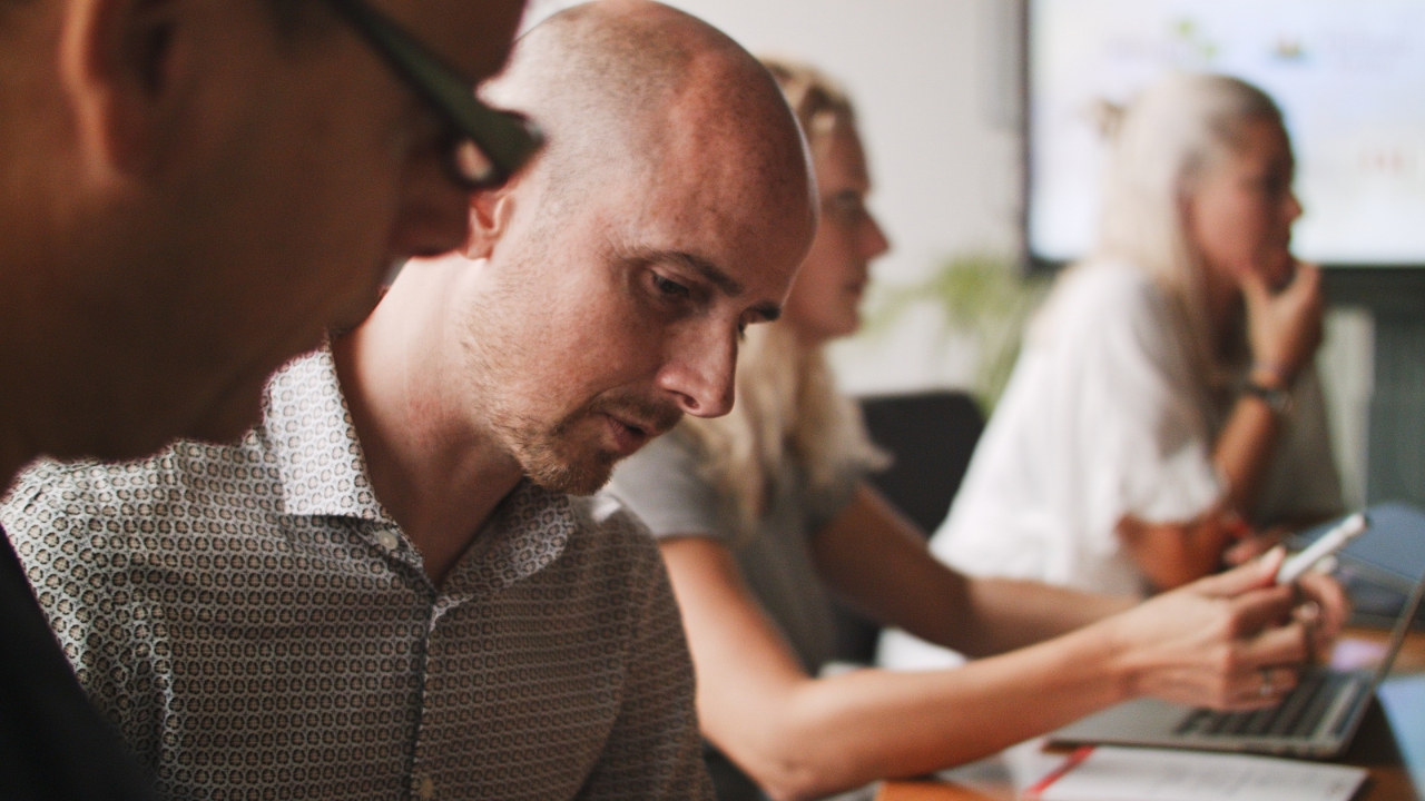 Still van de video. Man in gesprek met andere man. Twee vrouwen op de achtergrond